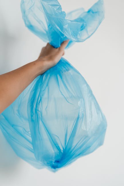 A person's hand holding a blue plastic rubbish bag, which appears to be filled with waste, against a plain white background. The bag is semi-transparent, showing some contents inside, and is gathered 